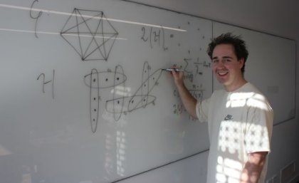 A man stands at a whiteboard, writing a mathematical equation. He is smiling at the camera.